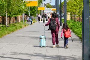 A mother and daughter walking with luggage outside Eindhoven Airport on a sunny day.